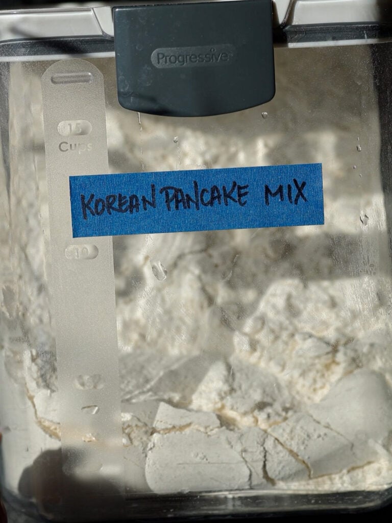A close-up of a plastic container labeled Korean Pancake Mix with blue tape. The container is filled with white powder and has a measurement strip on the side.